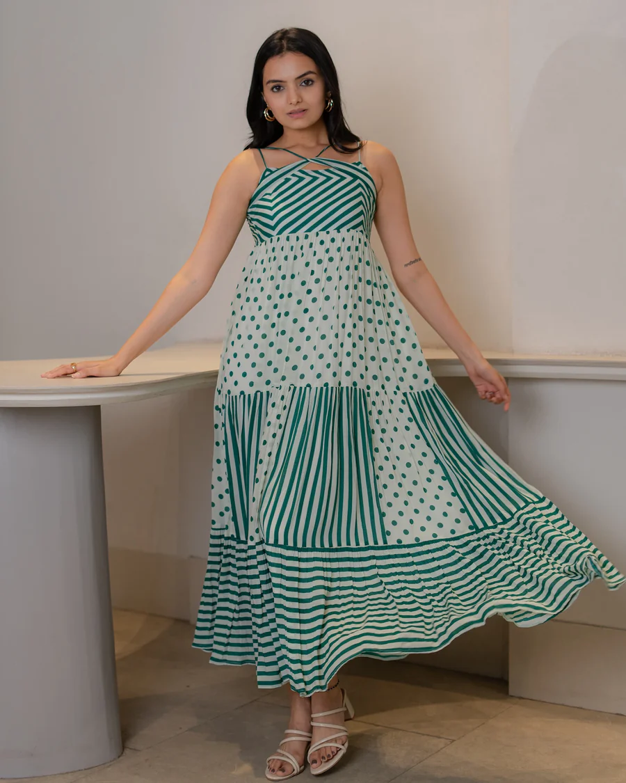 Woman wearing a sleeveless green and white A-line midi dress with polka dot and stripe patterns, standing indoors by a curved table.