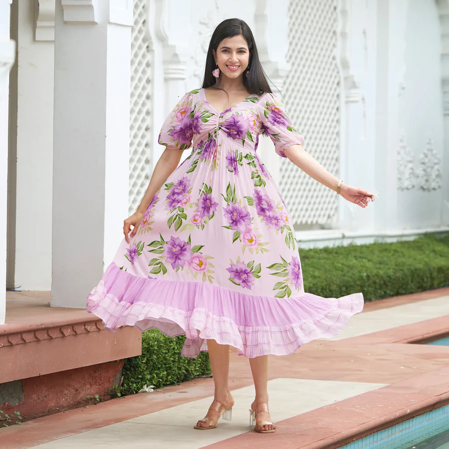 Woman wearing a pink floral A line midi dress with puff sleeves and ruffled hem, smiling and posing outdoors near a white architectural wall.